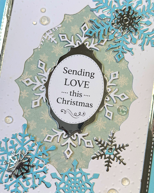 A close-up of a handmade Christmas card featuring layered scalloped backgrounds in mint green and white, embellished with die-cut snowflakes in light blue, white, and metallic silver. A central white oval contains the printed message 'Sending LOVE this Christmas'.
