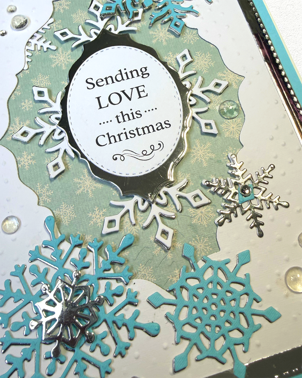 A close-up of a handmade Christmas card or craft plaque featuring layered die-cut snowflakes in white, silver, and turquoise, with a central white oval reading 'Sending LOVE .