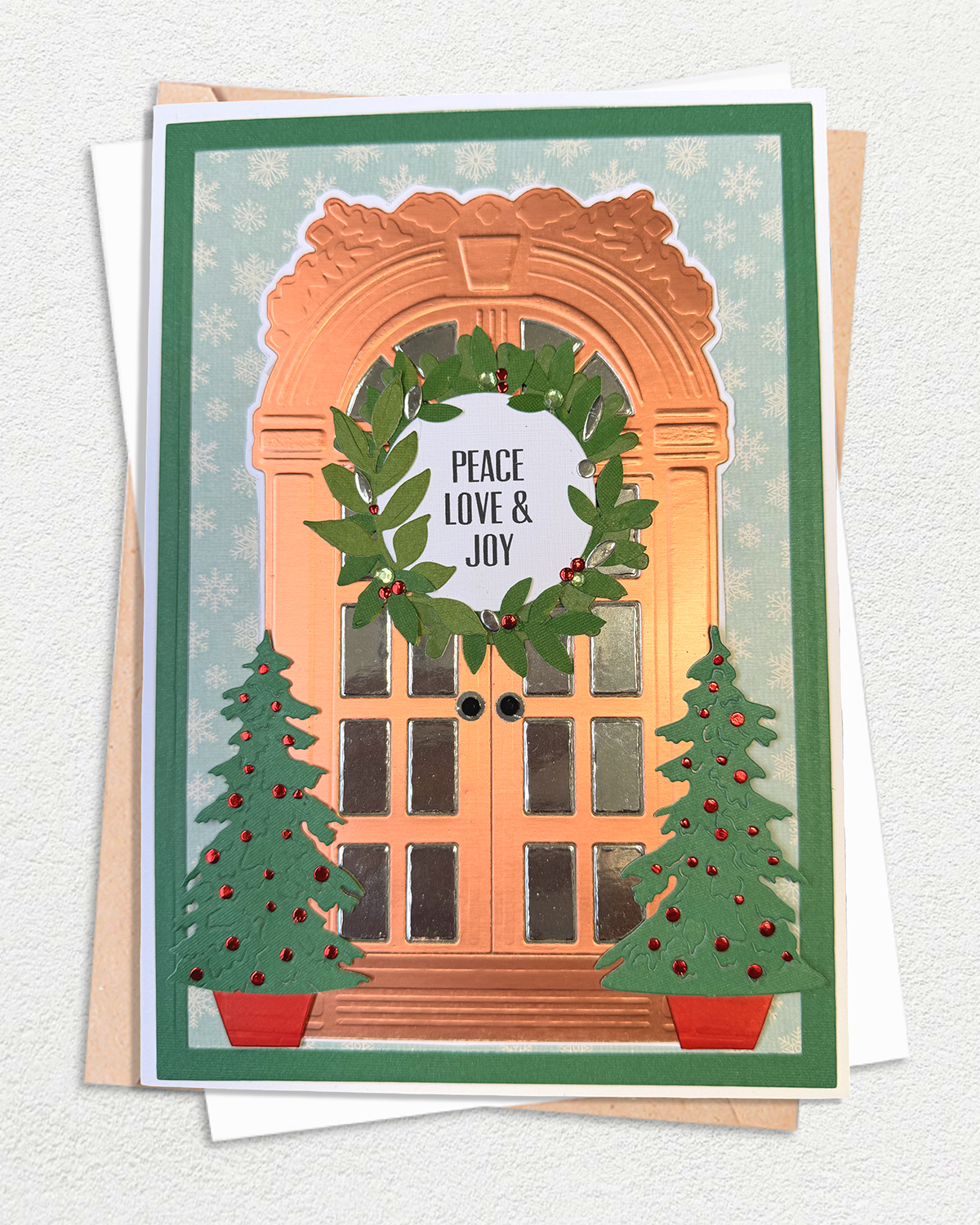 A handmade Christmas greeting card featuring an arched doorway with a green wreath that reads 'PEACE LOVE & JOY', flanked by two decorated Christmas trees, shown with envelopes on a textured white background.