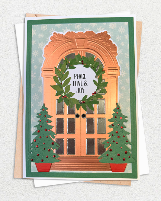 A handmade Christmas greeting card featuring an arched doorway with a green wreath that reads 'PEACE LOVE & JOY', flanked by two decorated Christmas trees, shown with envelopes on a textured white background.