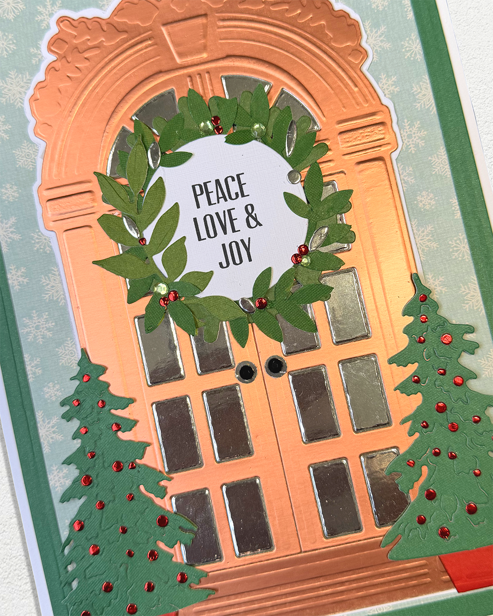 A close-up of a handmade holiday card featuring a copper-colored door with a green wreath that reads 'PEACE LOVE & JOY', decorated with two green Christmas trees and red ornaments, on a light blue snowflake background.