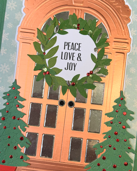 A close-up of a handmade holiday greeting card featuring a peach-colored door with silver window panes, a green leaf wreath with red berries, and the words 'PEACE LOVE & JOY' on a white oval center label. Two green Christmas trees with red ornaments decorate the lower corners.