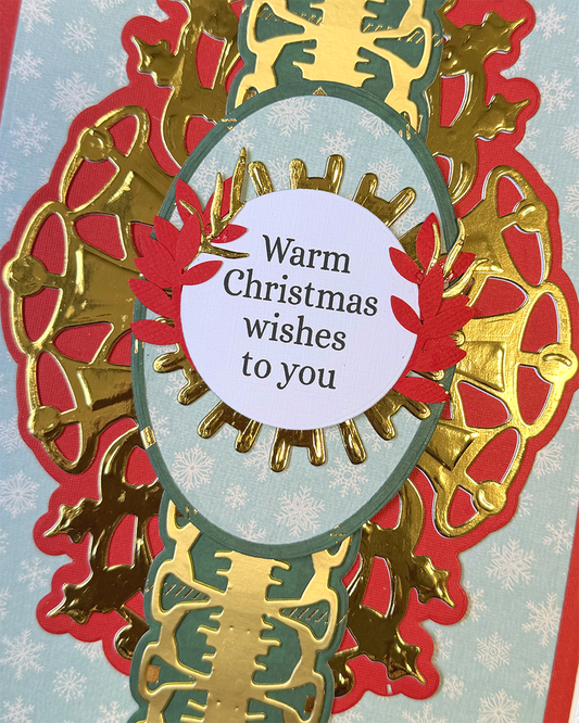 A close-up of a handmade Christmas greeting card featuring layered die-cut paper designs. The card has a light blue snowflake-patterned background, a large red and gold ornate frame, a central gold vertical bar, and a white oval sticker in the middle that reads 'Warm Christmas wishes to you'.