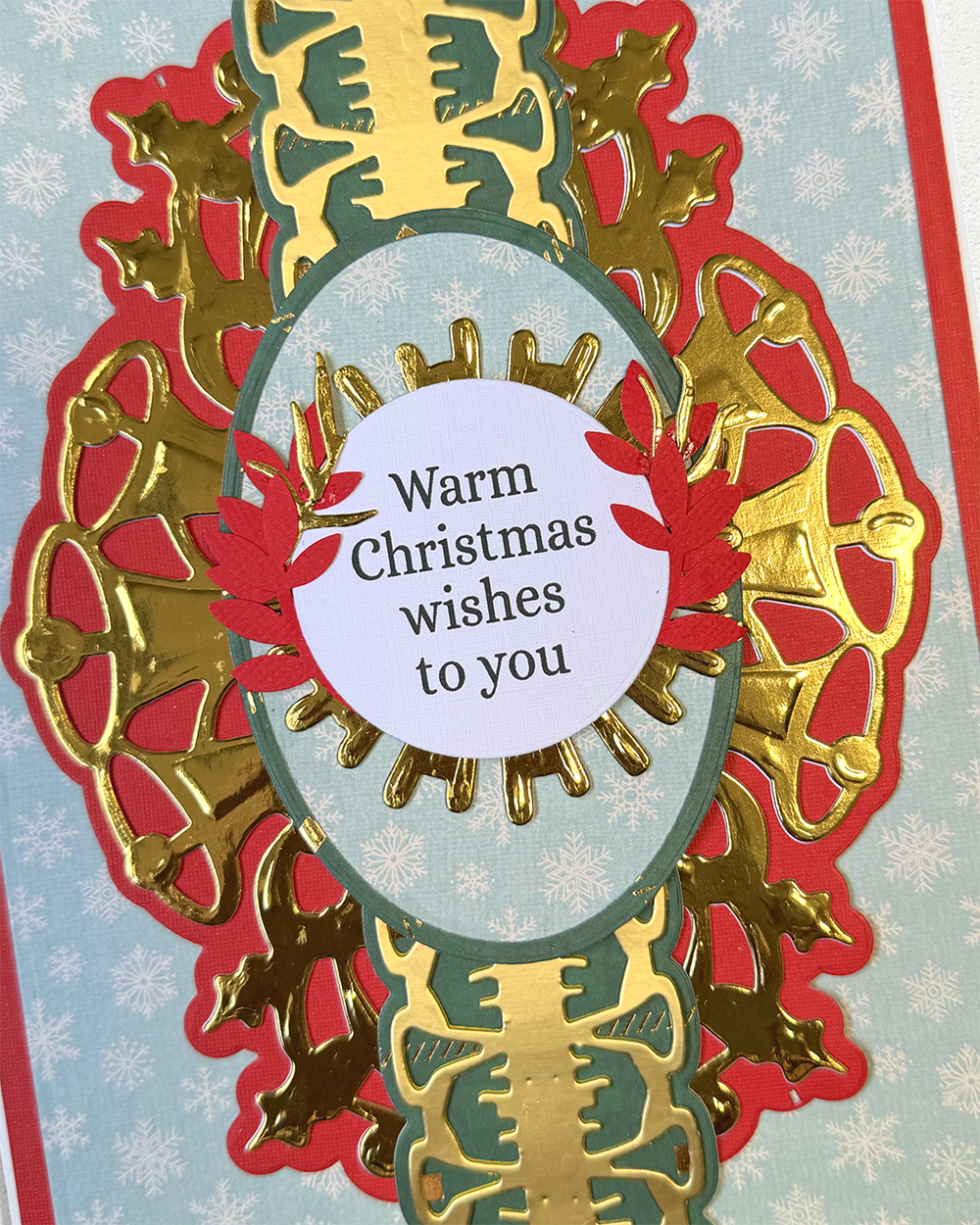 A close-up of a handmade Christmas greeting card featuring a layered design with red and gold scalloped borders, a light blue patterned background, and a central white oval with the text 'Warm Christmas wishes to you'.
