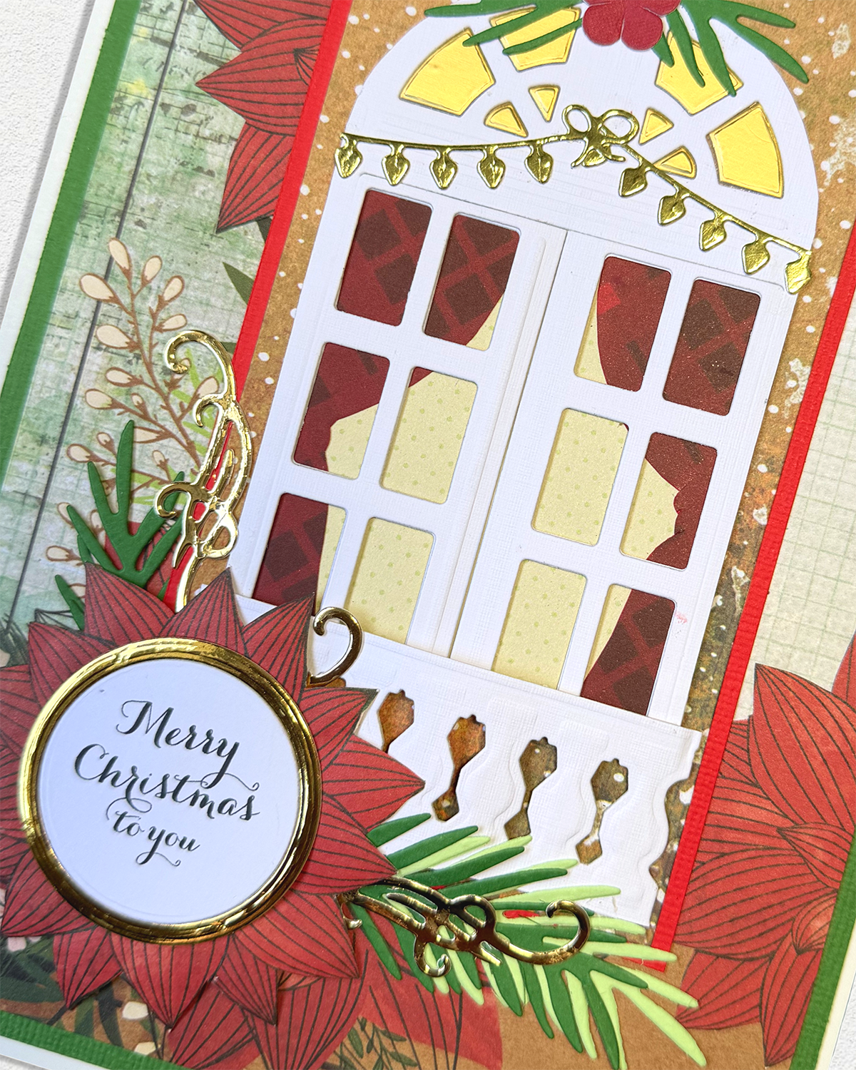 A close-up of a handmade Christmas card featuring a white arched window with red and gold patterned panes, surrounded by red poinsettias, green pine branches, gold ornaments, and a round white tag that reads 'Merry Christmas to you'.