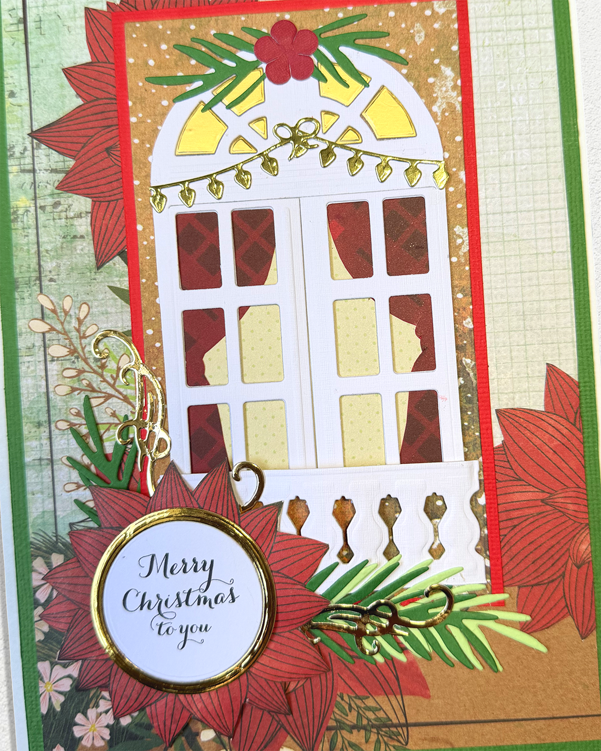 A handmade Christmas greeting card featuring a white window cutout with red and gold patterned panes, red poinsettia petals, green pine leaves, gold glitter accents, and a round badge that reads 'Merry Christmas to you'.