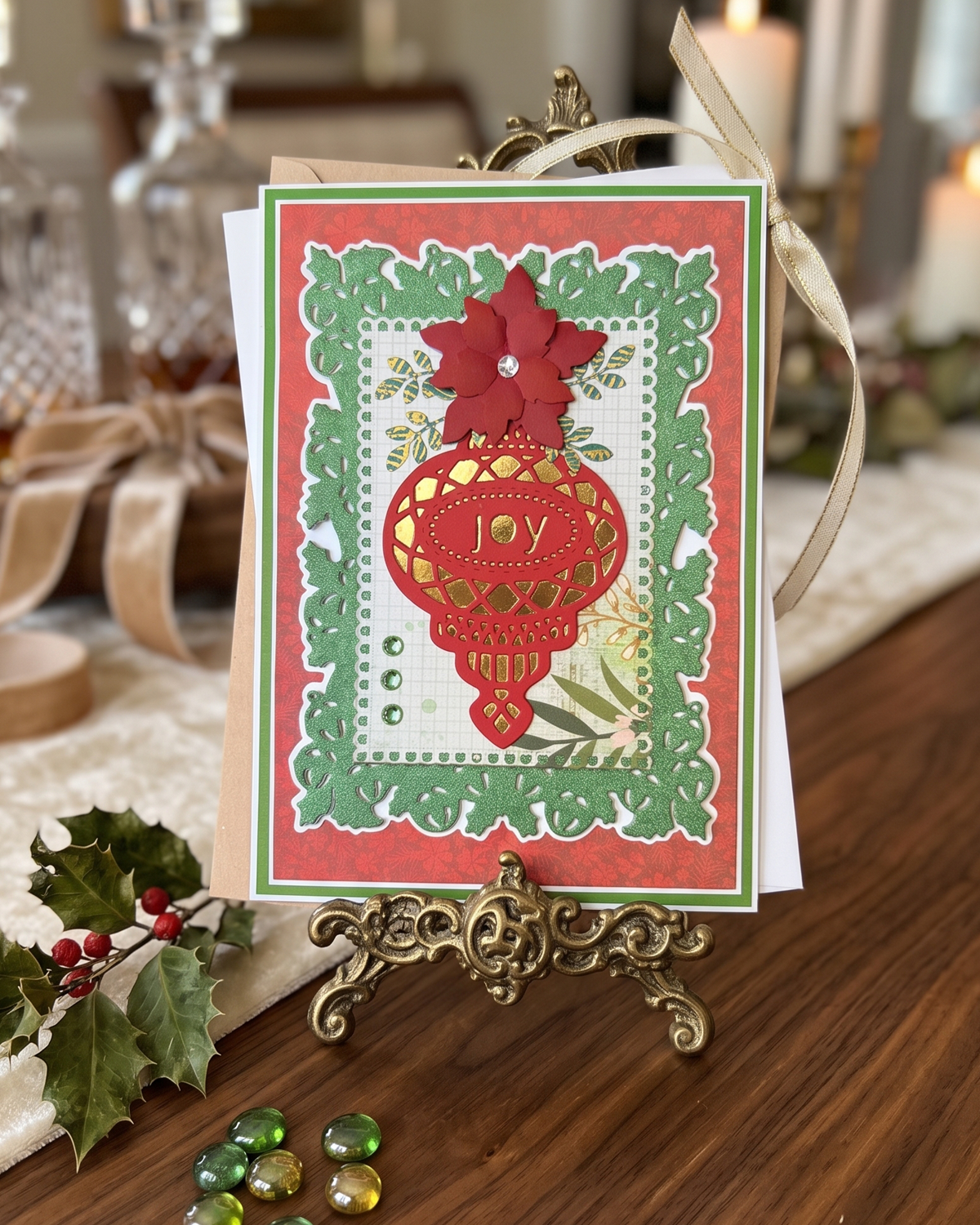 A handmade Christmas greeting card displayed on a small ornate gold stand on a wooden table. The card features a red hot air balloon, a red poinsettia, and green leafy borders on a red and white background. Made in NZ by Del Cards NZ.