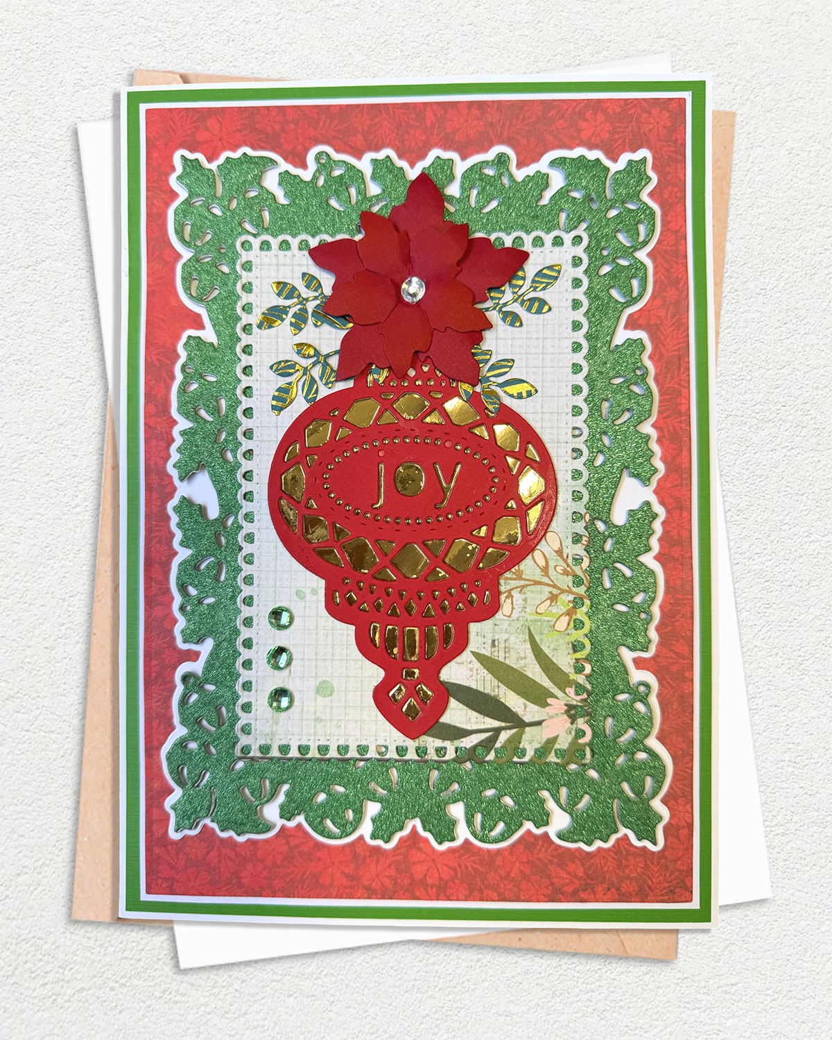 A handmade layered greeting card featuring a red poinsettia flower, a red lantern ornament with the word 'joy', and green leafy borders on a textured red background, displayed with white and kraft envelopes.