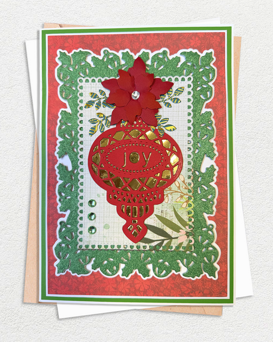 A handmade layered greeting card featuring a red poinsettia flower, a red lantern ornament with the word 'joy', and green leafy borders on a textured red background, displayed with white and kraft envelopes.