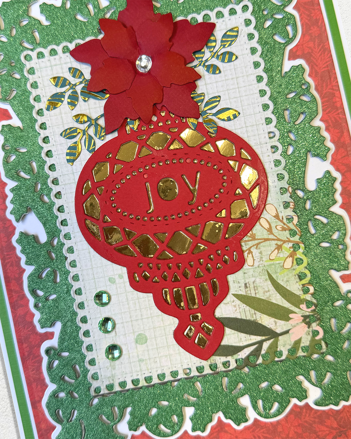 A close-up of a handmade layered paper card featuring a red hot air balloon with gold foil details and the word 'JOY' cut out in the center, topped with a red flower. The card is decorated with green glitter borders, a white grid background, and gold and green patterned paper accents.