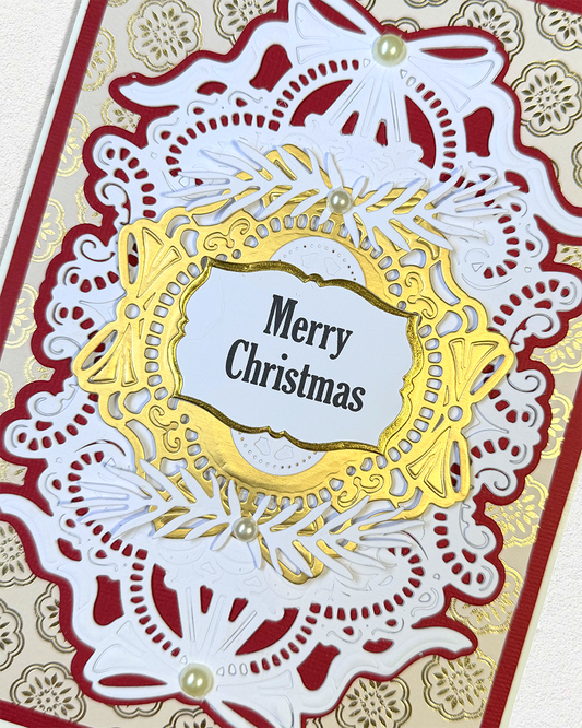 A close-up of an intricate handmade Christmas card featuring layered white and red laser-cut paper designs, gold foil accents, and a central white label that reads 'Merry Christmas'.