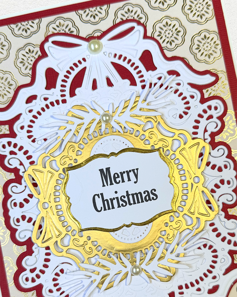 A close-up of a handmade Christmas card featuring intricate layered die-cut paper designs in red, white, and gold, with a central white label reading 'Merry Christmas' and small pearl embellishments.