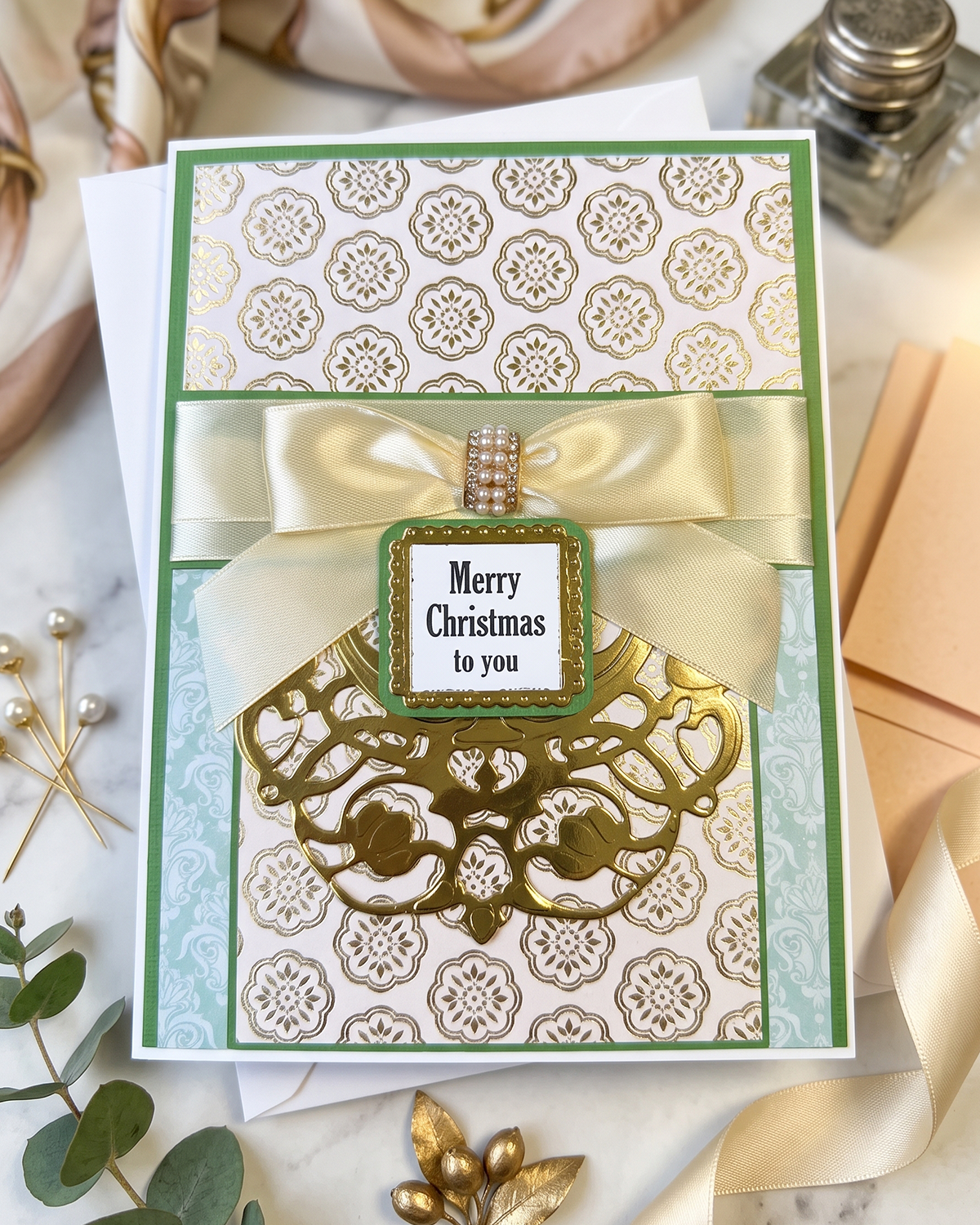 A handmade Christmas greeting card with a green and gold patterned background, a cream satin ribbon bow, and a central label that reads 'Merry Christmas to you.' The card is styled on a marble surface with decorative gold leaves, a peach envelope, and a small bottle in the background.