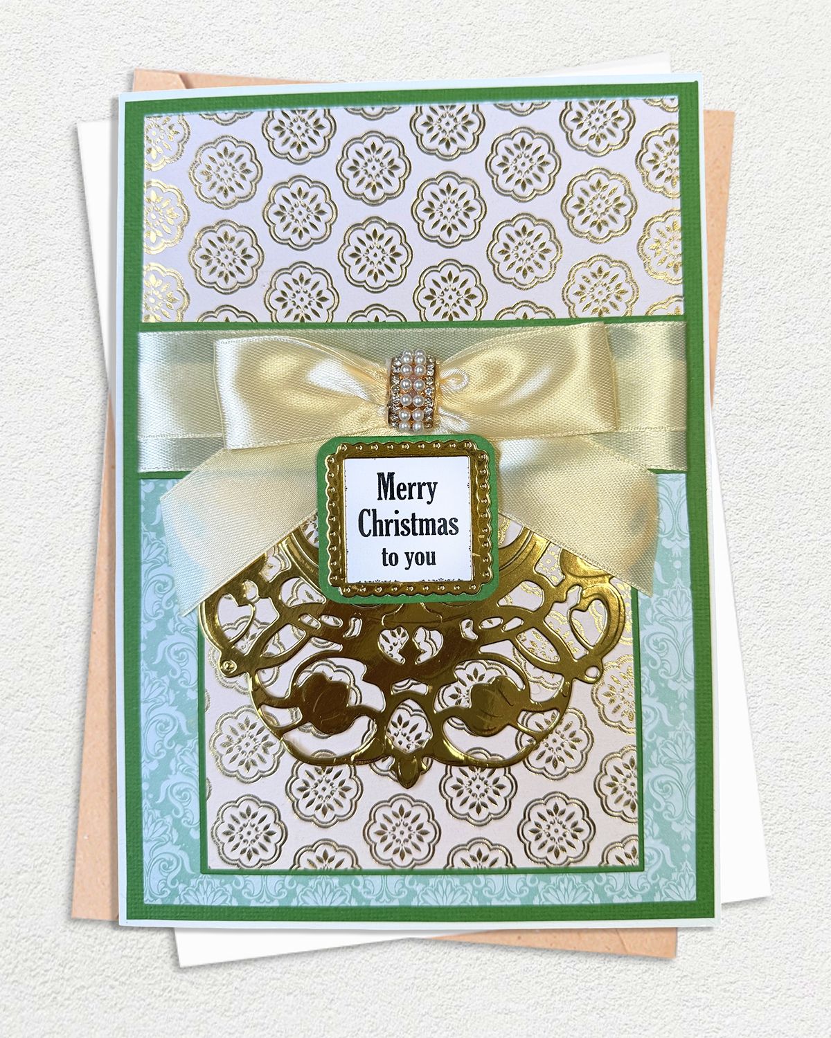 A handmade Christmas greeting card with a green border, gold patterned paper, a cream satin bow, and a central label reading 'Merry Christmas to you', displayed on top of other cards against a light textured background.