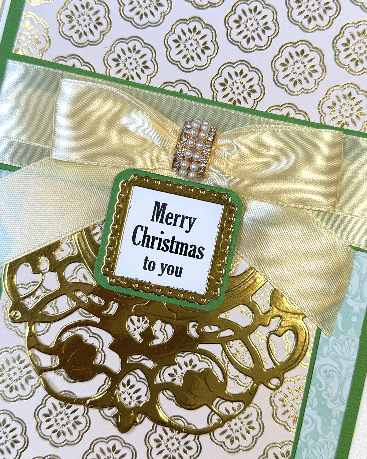 A close-up of a handmade Christmas greeting card featuring a large cream satin bow with a rhinestone-embellished center, a green and gold laser-cut ornament, and a central white tag that reads 'Merry Christmas to you'.