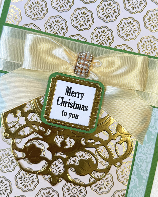 A close-up of a handmade Christmas greeting card featuring a large cream satin bow with a rhinestone-embellished center, a green and gold laser-cut ornament, and a central white tag that reads 'Merry Christmas to you'.