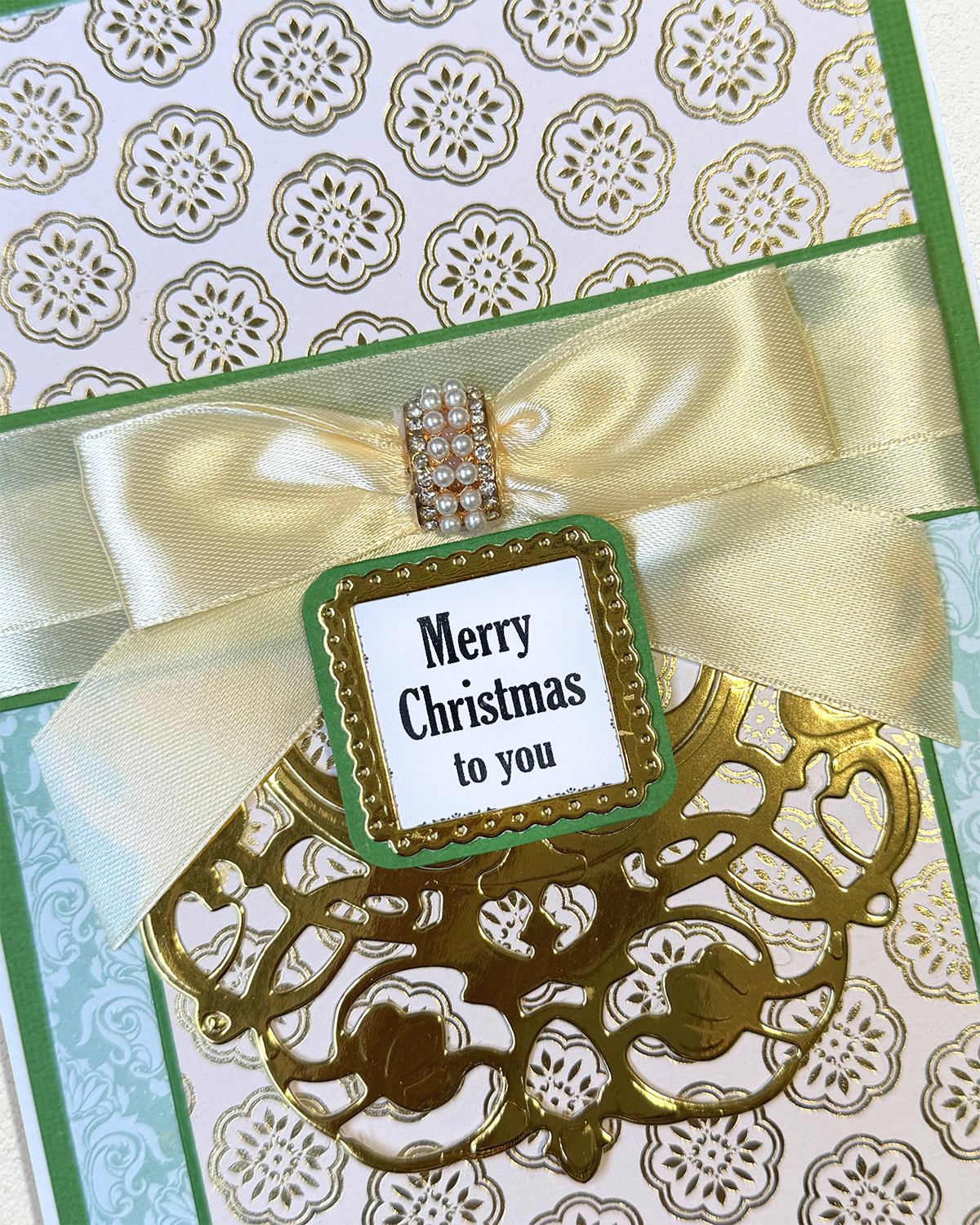 A close-up of a handmade Christmas greeting card featuring a gold laser-cut filigree design, a green and gold patterned background, a cream satin ribbon bow, and a central tag that reads 'Merry Christmas to you'.