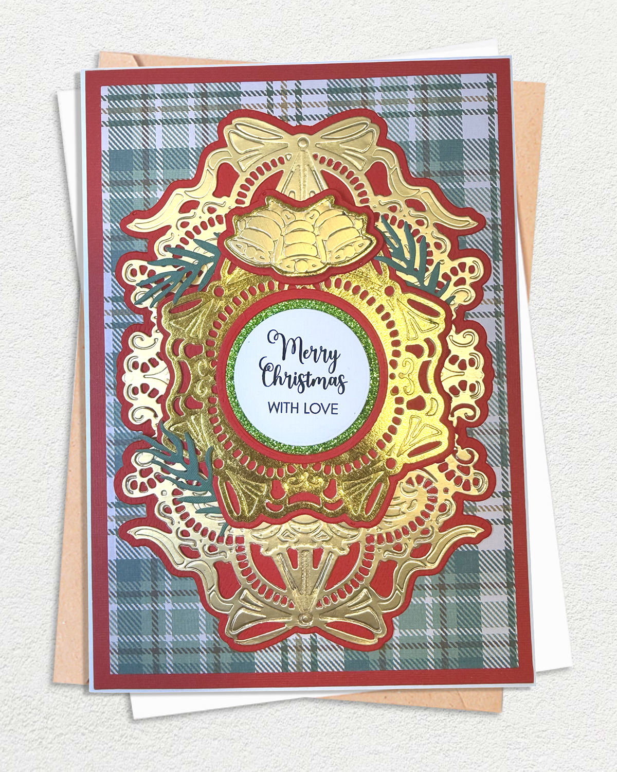 A handmade Christmas card featuring an ornate gold and red scalloped frame with a central white circle that reads 'Merry Christmas with Love,' layered over a blue and green plaid background. The card is shown with kraft paper envelopes behind it on a light textured surface.