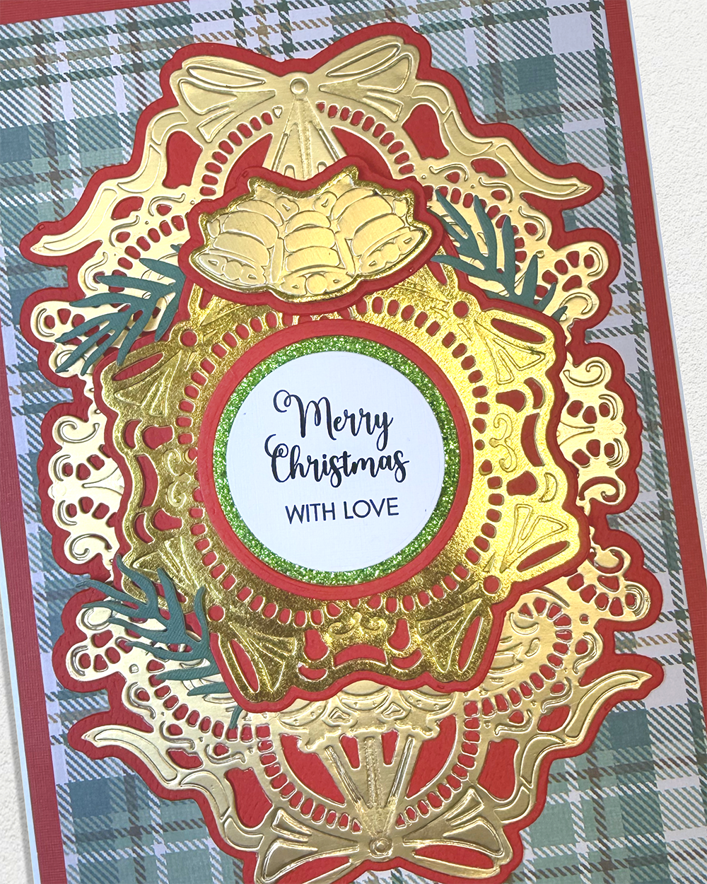 A close-up of a handmade Christmas card featuring an intricate layered die-cut design in gold, red, and green, with a central white circle that reads 'Merry Christmas WITH LOVE'. The card is layered over a green and white plaid background.