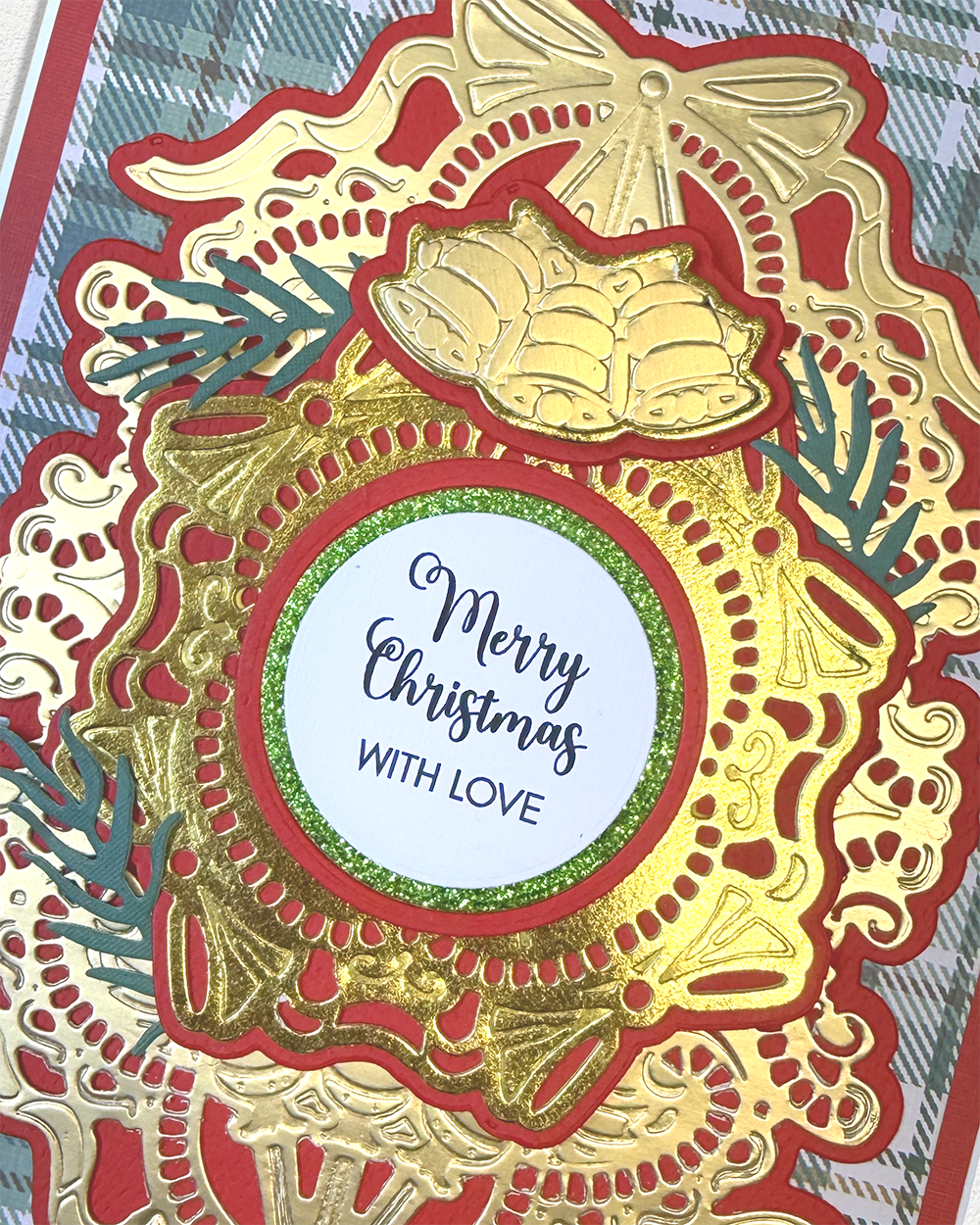 A close-up of a handmade Christmas card featuring an intricate layered design with gold and red die-cut embellishments, a glittery green border, and a central white circle that reads 'Merry Christmas WITH LOVE'.