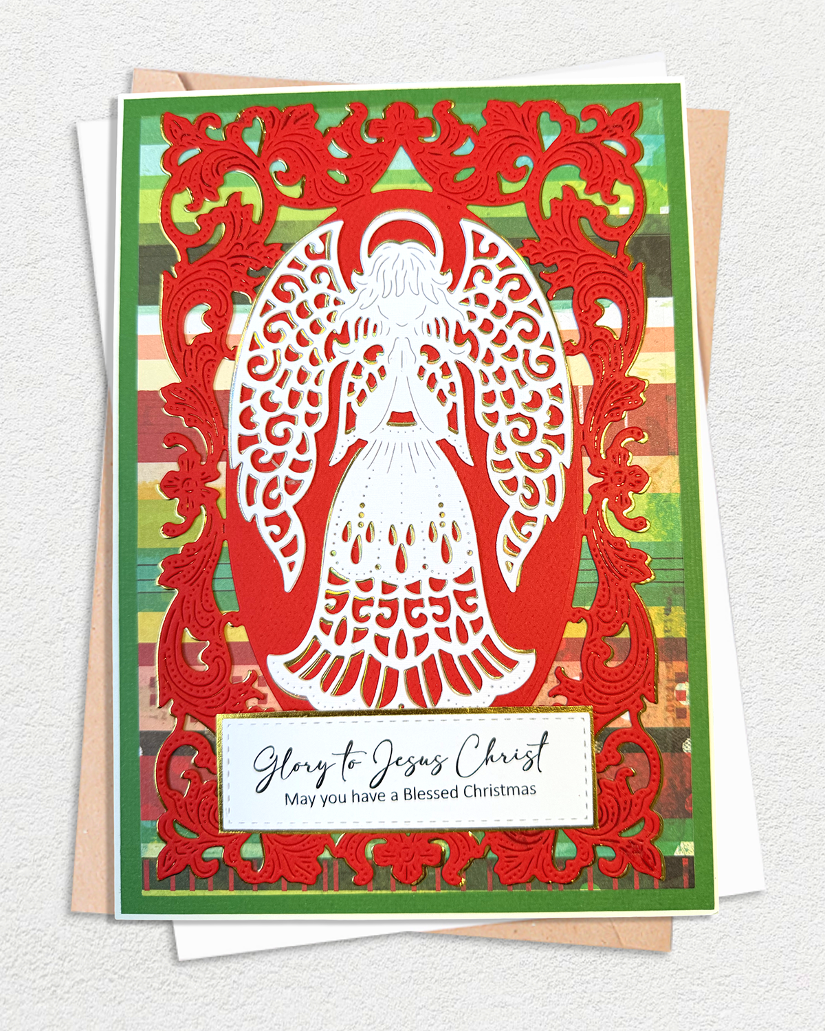 A layered Christmas greeting card featuring a white angel silhouette cutout over a red ornate border, with a green striped background and a white message panel reading 'Glow to Jesus Christ May you have a Blessed Christmas.' The card is photographed on top of white envelopes against a light textured wall.