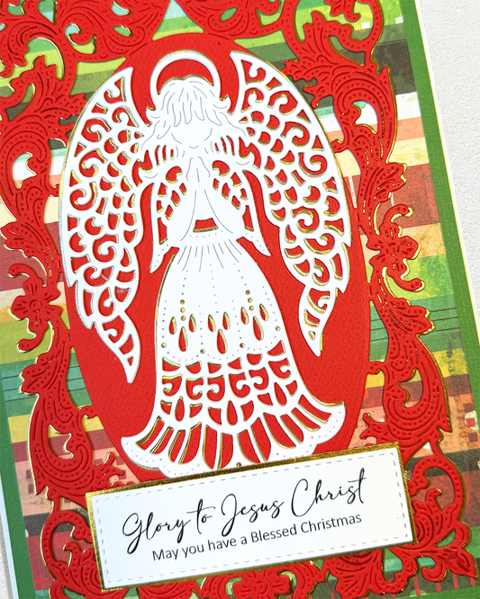 A close-up of a handmade Christmas card featuring a white angel silhouette layered over a red ornate frame, with a green patterned background and a white label that reads, 'Glory to Jesus Christ May you have a Blessed Christmas.'.