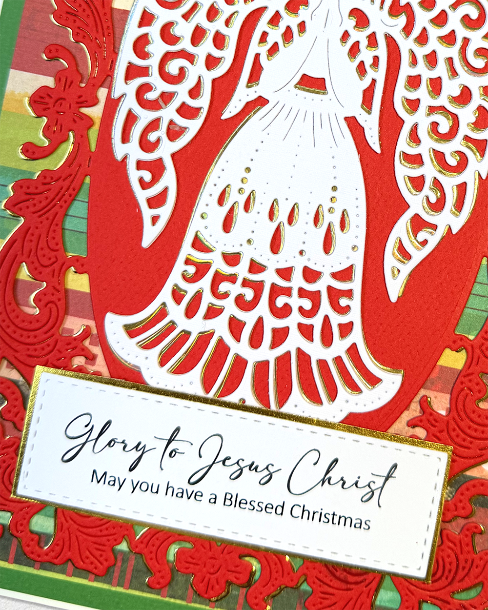 A close-up of a handmade Christmas greeting card featuring a white angel silhouette cutout over a red oval, with red ornate border details and a white banner that reads, 'Glory to Jesus Christ May you have a Blessed Christmas.'.