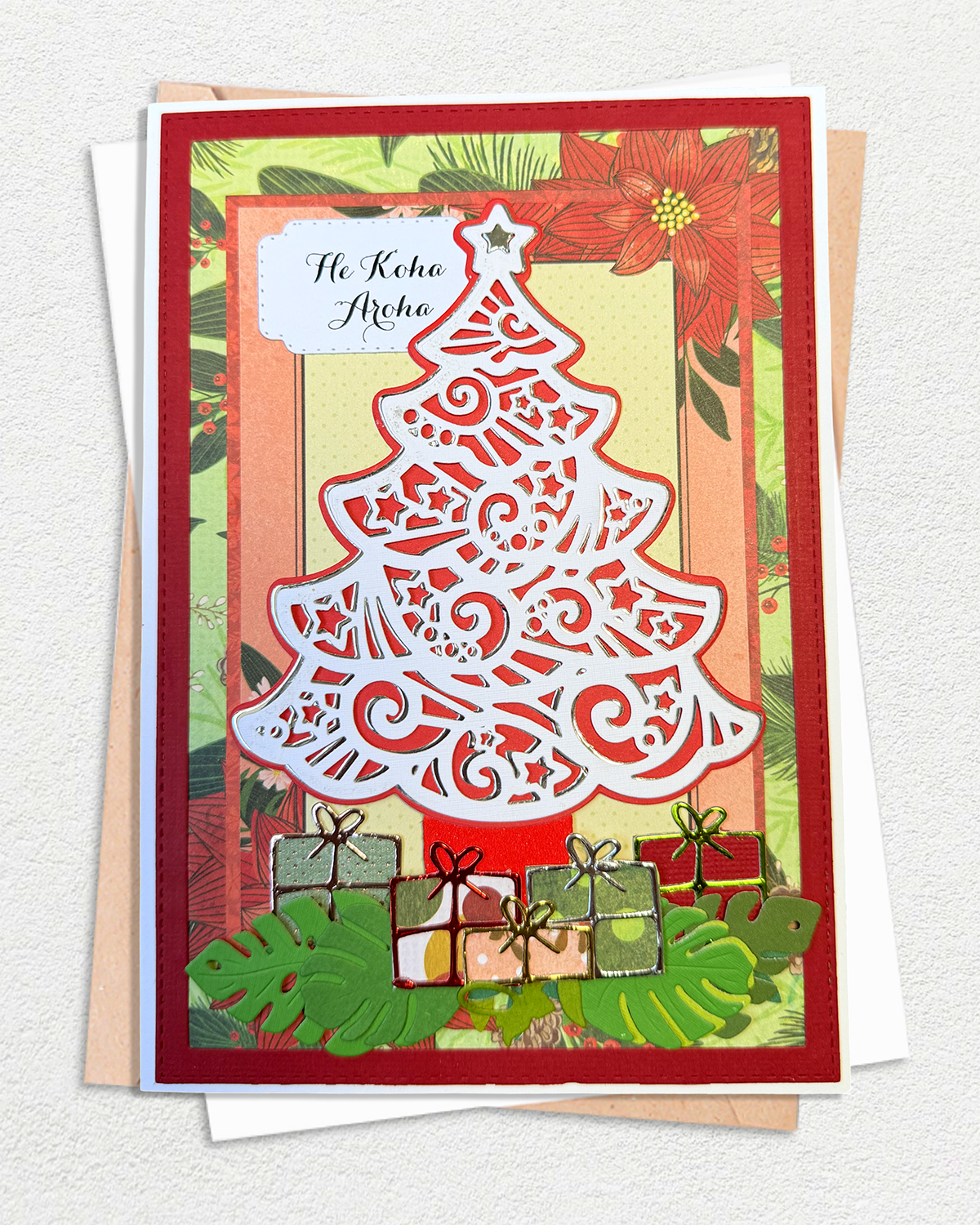 A handmade Christmas greeting card featuring a white laser-cut tree design, red poinsettias, green leaves, and wrapped gifts at the bottom, displayed on top of layered envelopes against a light textured background.