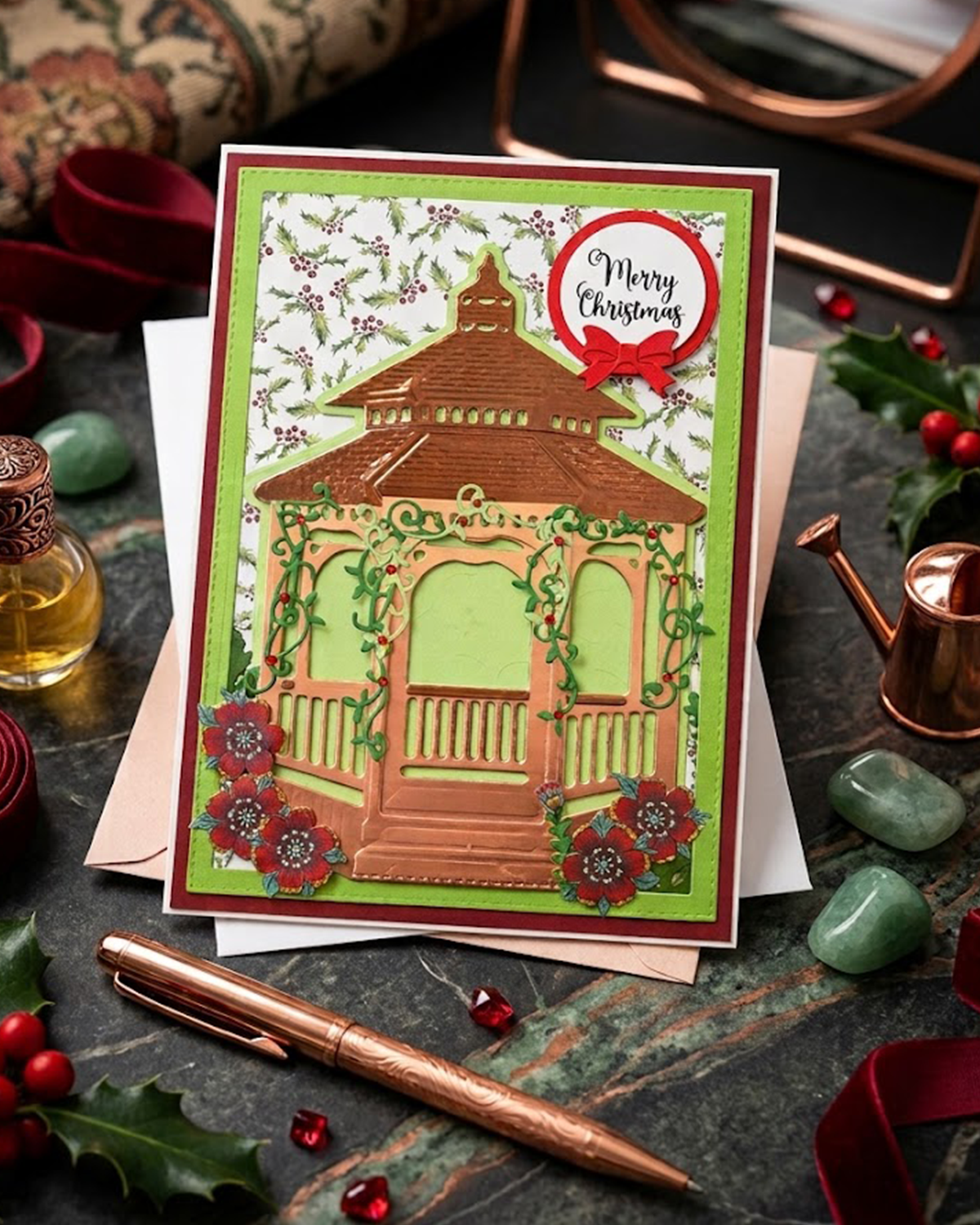 A handmade Christmas greeting card featuring a 3D layered gazebo design with green vines, red flowers, and a 'Merry Christmas' tag, styled on a dark stone surface with holiday props.