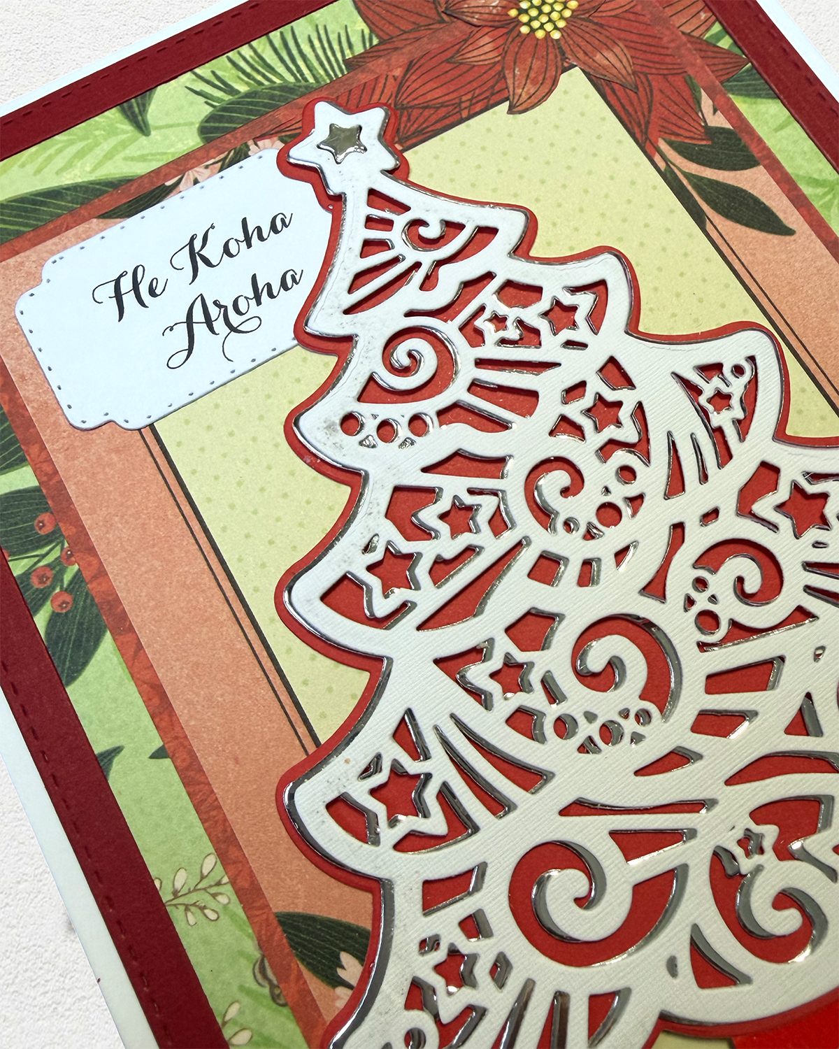 A close-up of a handmade Christmas card featuring a large white laser-cut tree design with red layered borders, a green patterned background, and a white label that reads 'He Koha Aroha'.