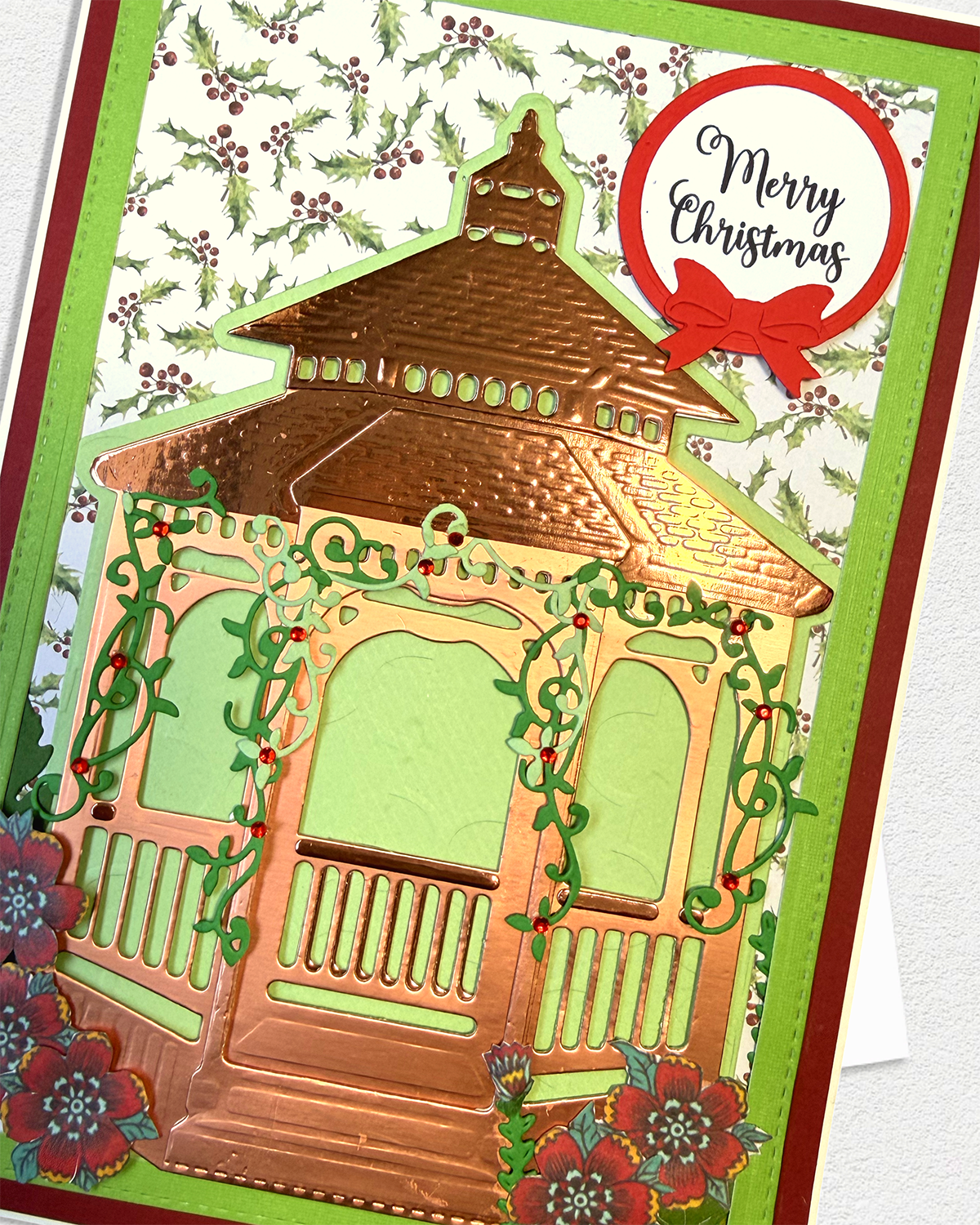 A handmade Christmas greeting card featuring a die-cut copper-toned gazebo with green vine details, red flowers, a holly-patterned background, and a red circular 'Merry Christmas' badge.