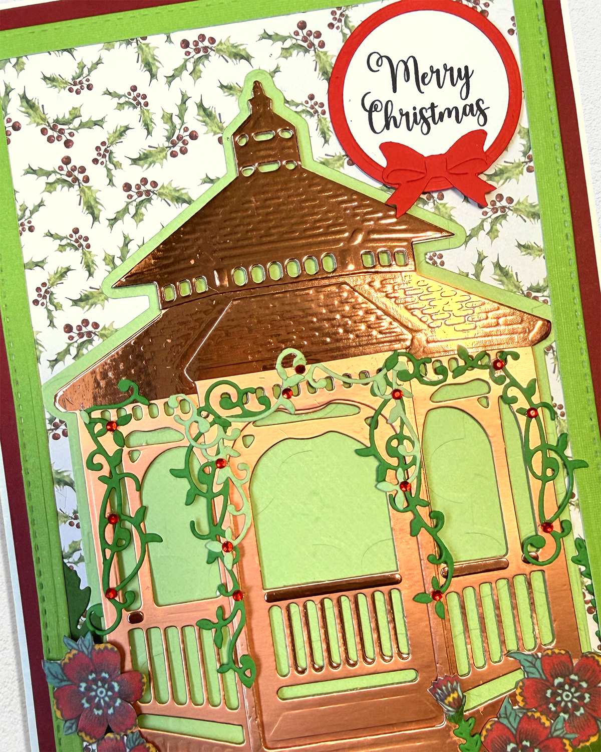A close-up of a handmade Christmas greeting card featuring a die-cut copper-toned gazebo with green vine accents, a red 'Merry Christmas' badge with a bow, and a holly-patterned background.