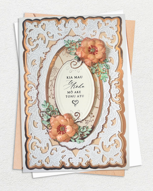 An ornate layered greeting card with a white laser-cut floral border, tan and brown inner frames, and raised peach flowers with green leaves. The center reads 'KIA MAU Sheha Mō Ake Tōnu Atu' in black script and block lettering. Maori wedding card for sale by Del Cards NZ.