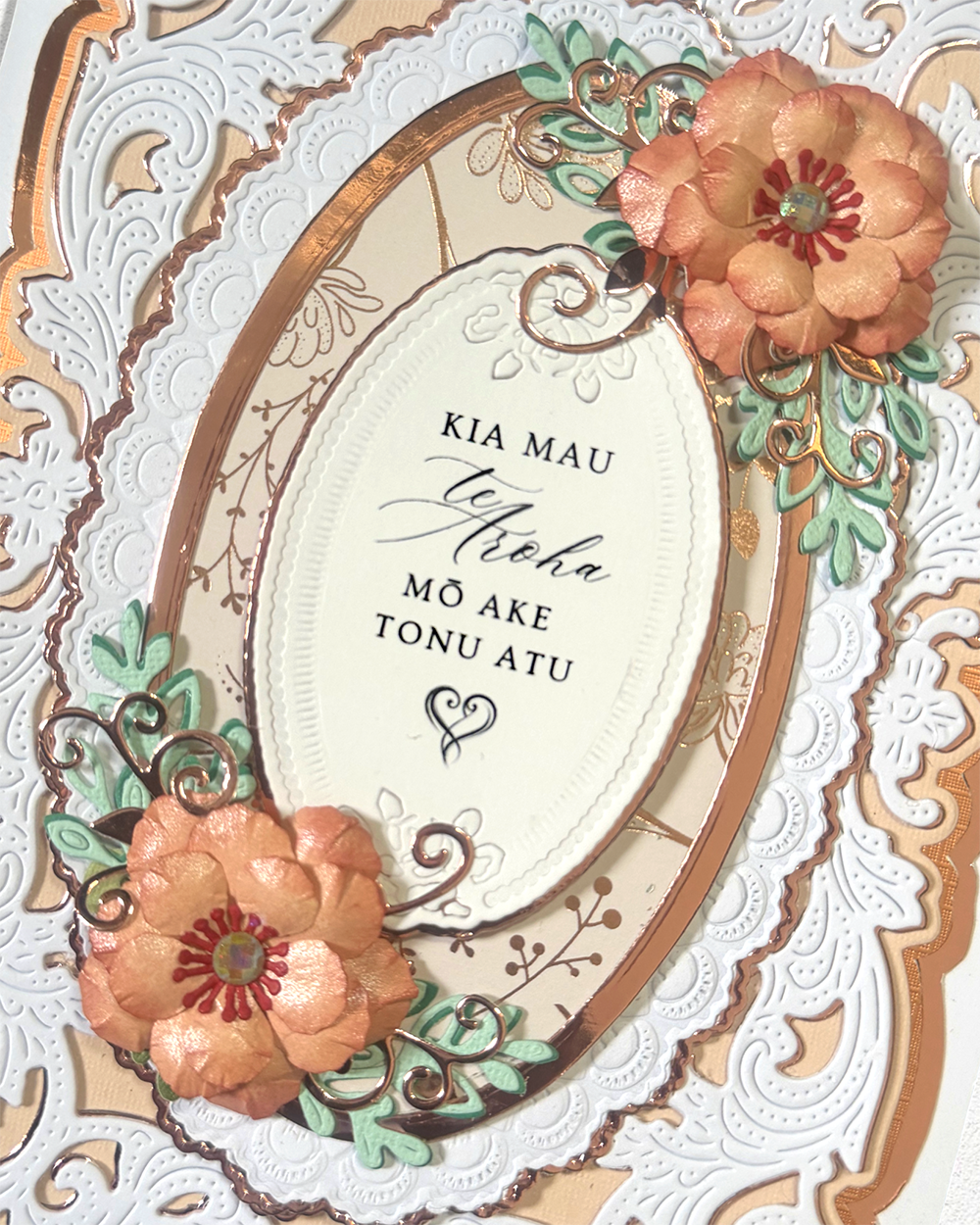 A close-up of a decorative floral card or art print featuring an ornate white embossed border, a cream central oval, copper-toned framing, and two peach-pink layered flowers with green leaves and small gold accents. The center contains the text 'KIA MAU te Nihoa MÔ AKE TONU ATU' in black script and serif lettering.