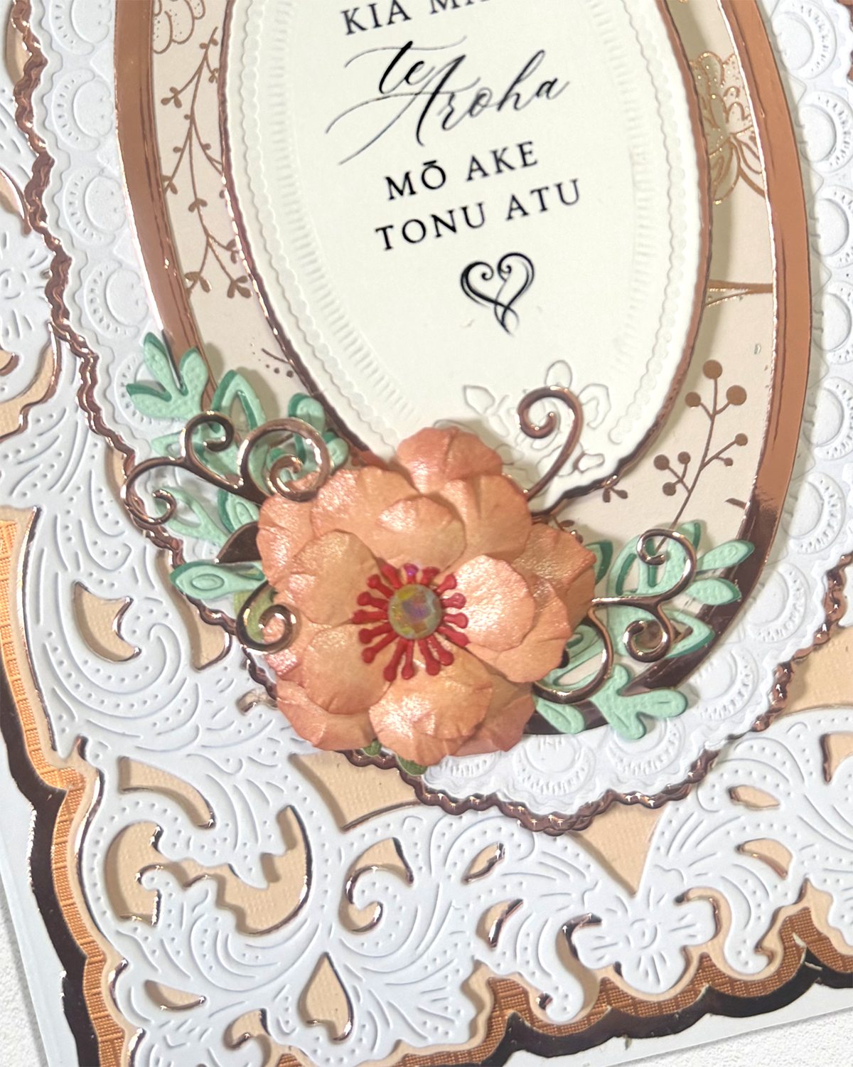 Close-up of a decorative handmade card featuring layered oval frames, embossed floral details, and a central peach flower with green leaves and a red center, with a cream oval insert containing Māori and English text.
