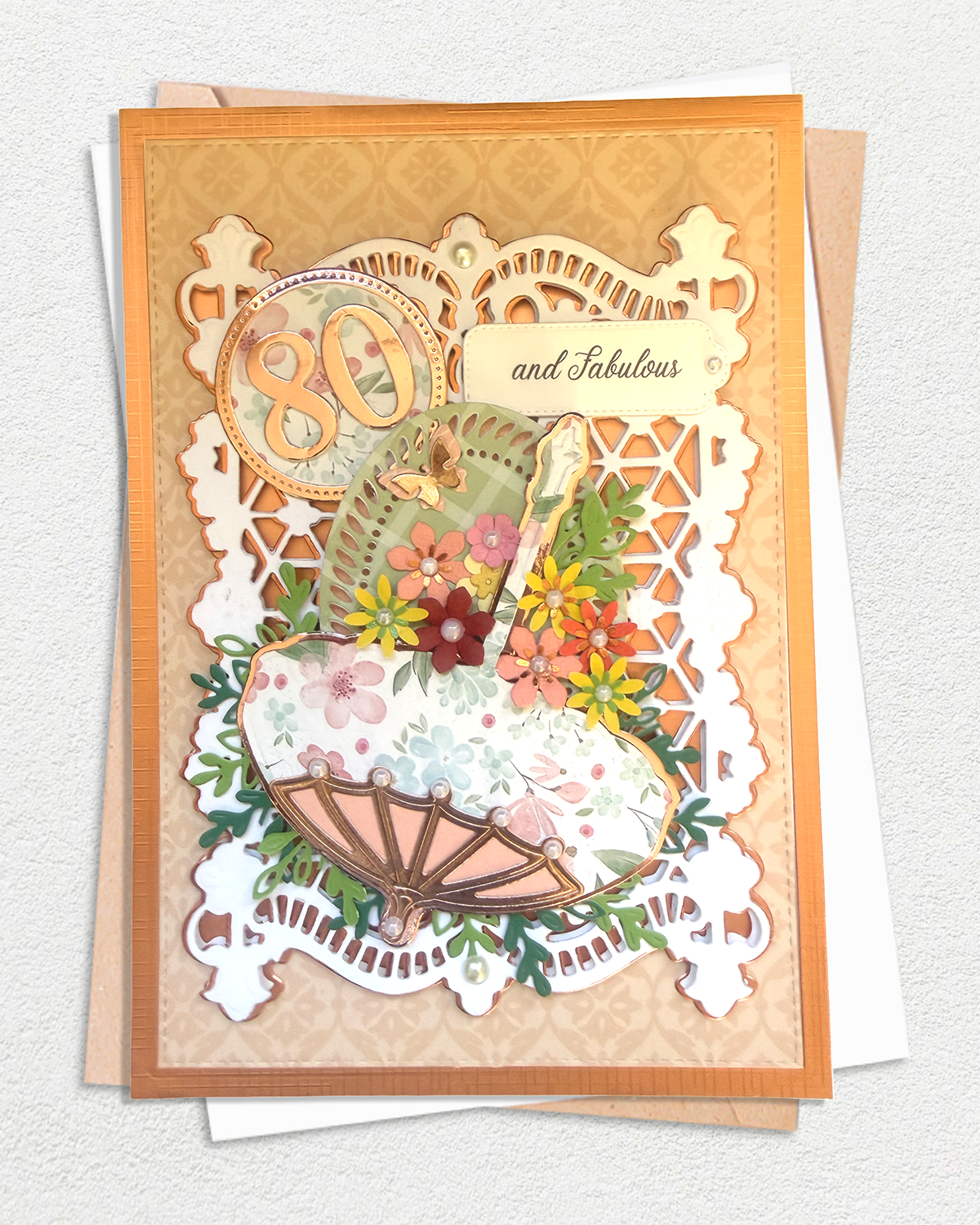 An elegant layered card with a vintage-inspired 80th birthday design, featuring a floral patterned hat, a folding umbrella, flowers, and a scalloped white lace overlay with the words '80 and Fabulous'.