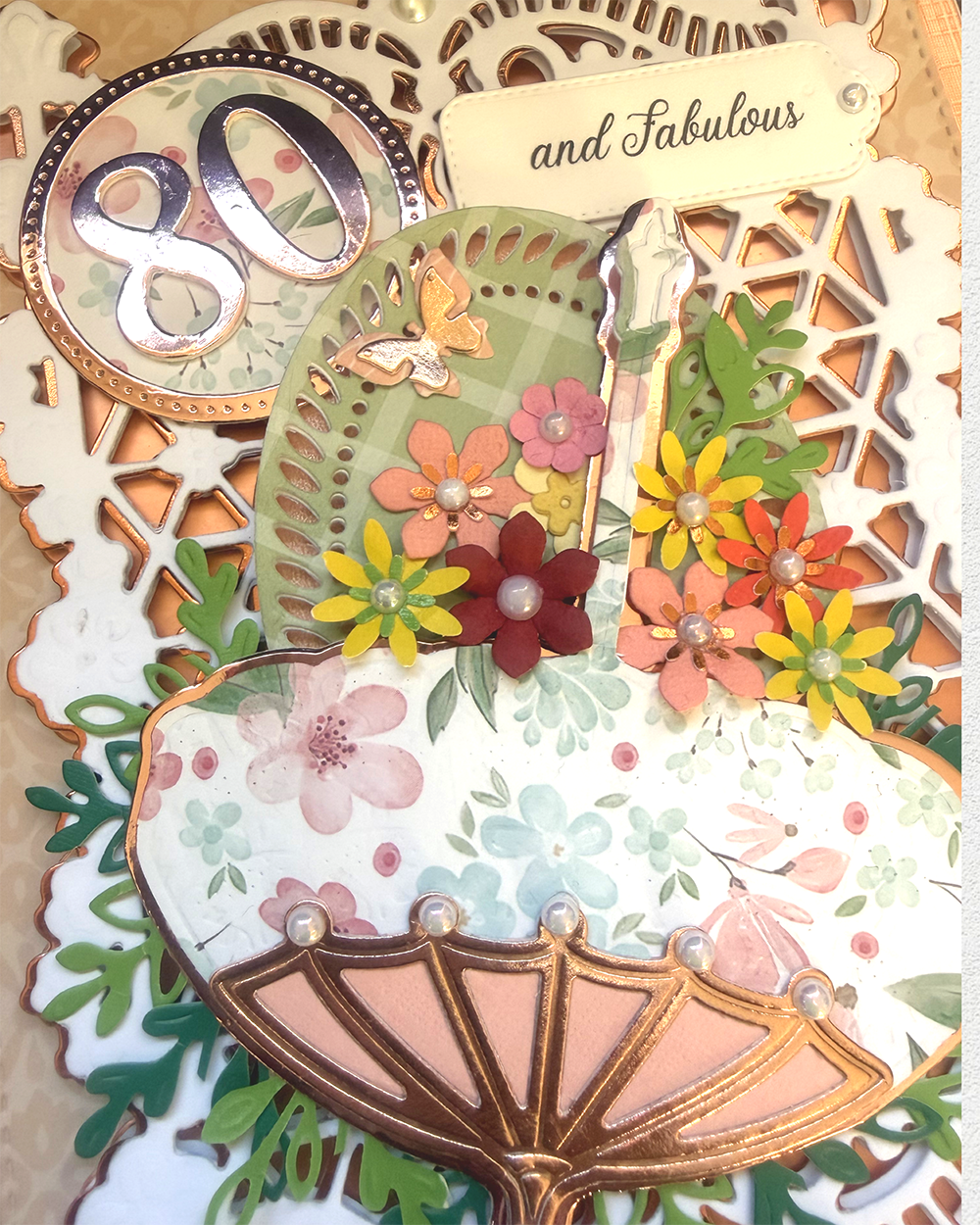 A close-up of a decorative handmade card or scrapbook page featuring layered paper elements, including a scalloped fan shape, floral cutouts, a butterfly embellishment, a small candle, and a tag reading 'and Fabulous.' The design uses many pastel and bright colors with metallic gold accents, so the overall color identity is multicolor rather than one dominant simple hue.