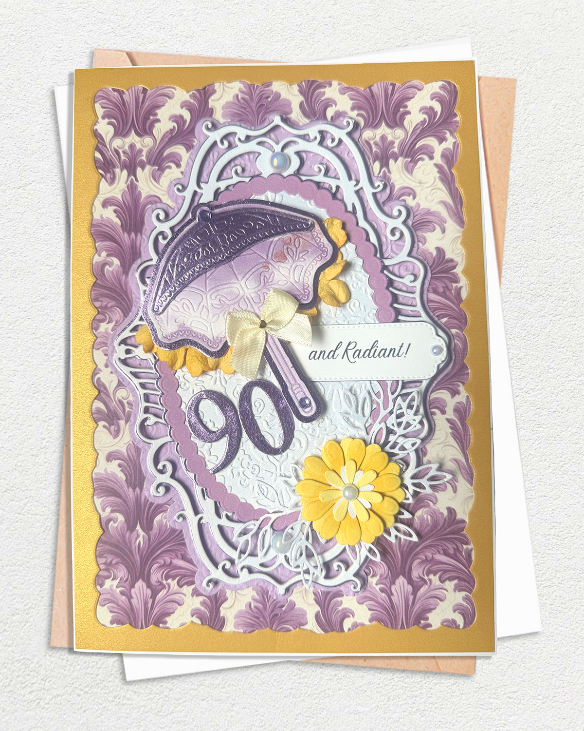 A 90th birthday card with a purple and lavender damask pattern, featuring a purple umbrella, yellow flowers, and the number 90 in raised lettering, displayed on top of kraft paper envelopes.