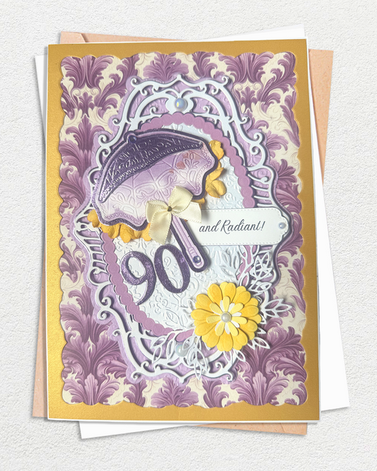 A 90th birthday card with a purple and lavender damask pattern, featuring a purple umbrella, yellow flowers, and the number 90 in raised lettering, displayed on top of kraft paper envelopes.