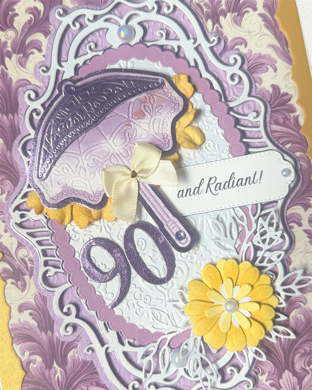 A close-up of a decorative handmade card featuring a lavender and purple embossed umbrella, yellow flower accents, white lace details, and the number 90 with the phrase 'and Radiant!!' on a patterned background.