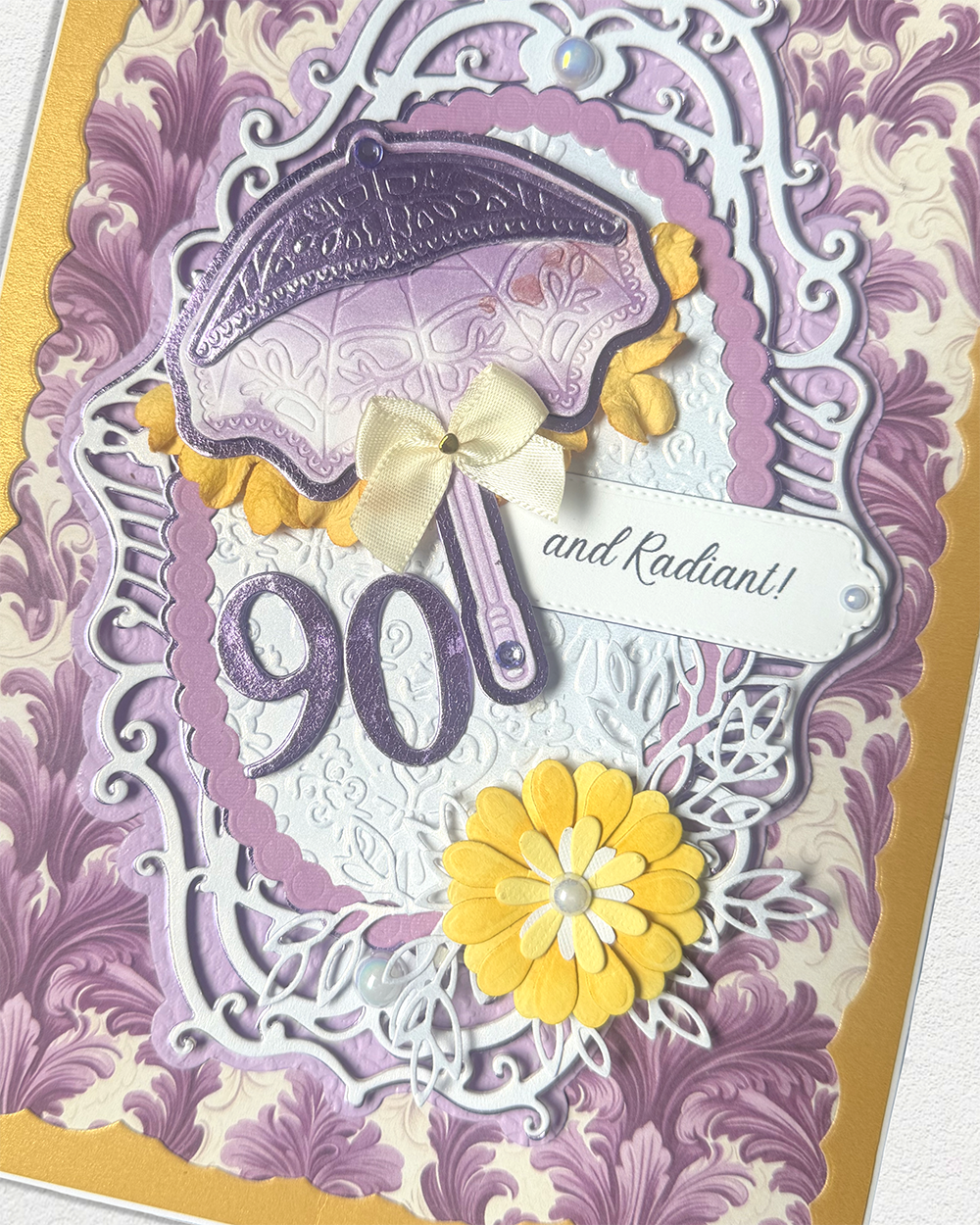 A close-up of a decorative handmade greeting card featuring a purple and lavender damask pattern, a purple umbrella, a yellow flower, and the number 90 with the phrase 'and Radiant!'.