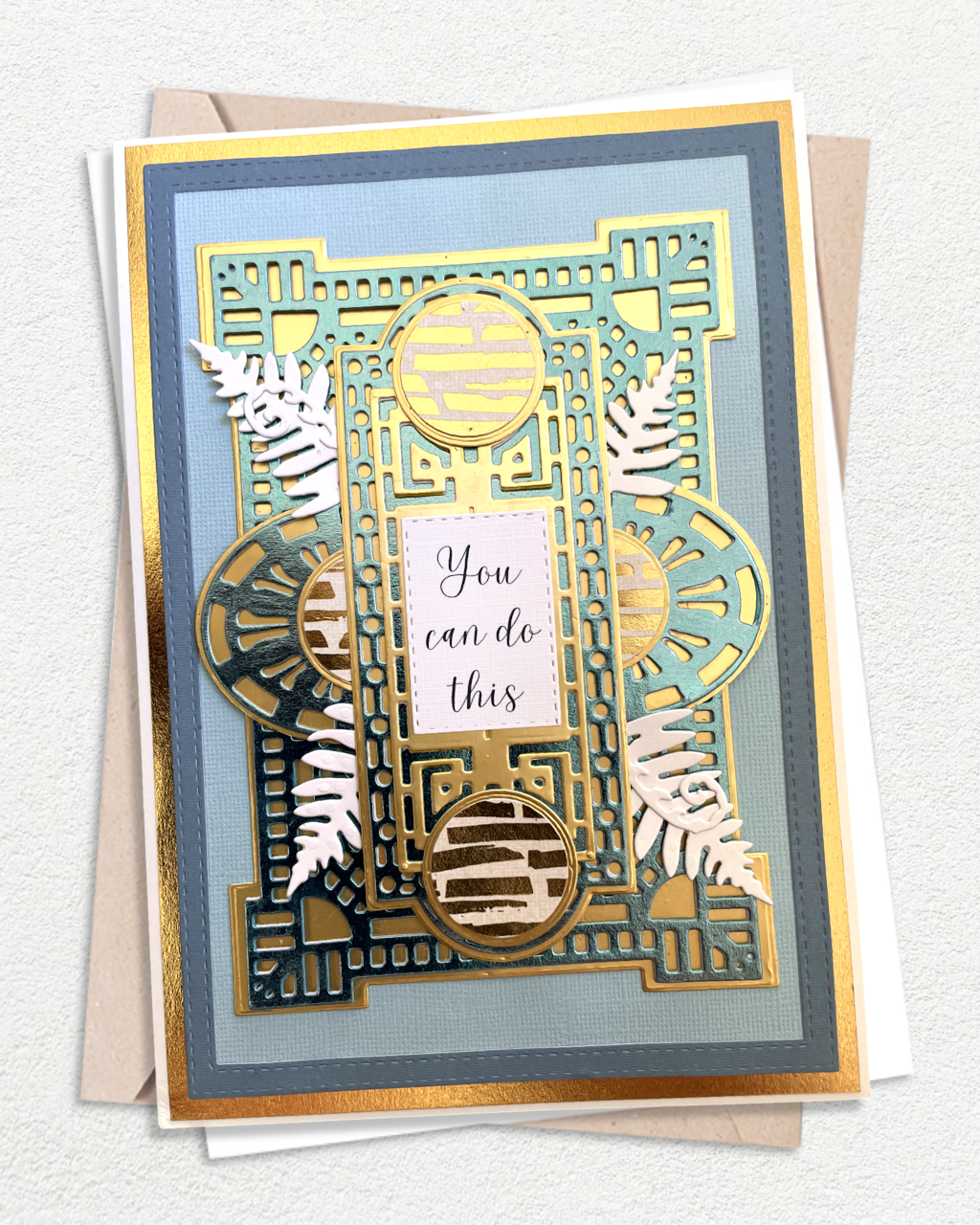 Handmade luxury encouragement card on a soft steel blue cardstock base with a gold foil mat. An intricate geometric gold die-cut architectural frame with Art Deco-style detailing spans the full card face, layered over a soft sage green panel. Two white silver fern fronds — a distinctly New Zealand motif — flank the centre. A circular gold element sits at the top, with a blush pink rectangular sentiment tag reading "You can do this" in handwritten script at the centre. Made by Del Cards.