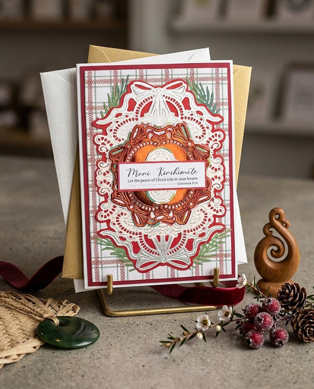 A handmade layered greeting card with a red and white plaid border, an intricate white lace overlay, and a central orange wreath design, displayed on a small stand with envelopes and autumn-themed props in the background.
