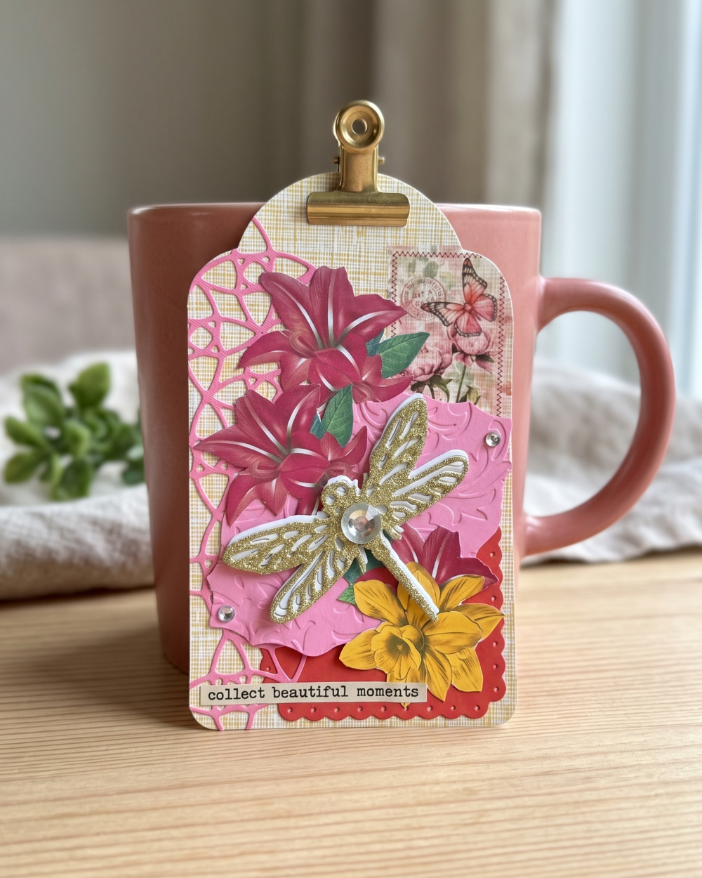 A decorative paper craft tag with layered floral designs, a dragonfly embellishment, and the phrase 'collect beautiful moments' at the bottom, clipped to a pink mug on a wooden table.