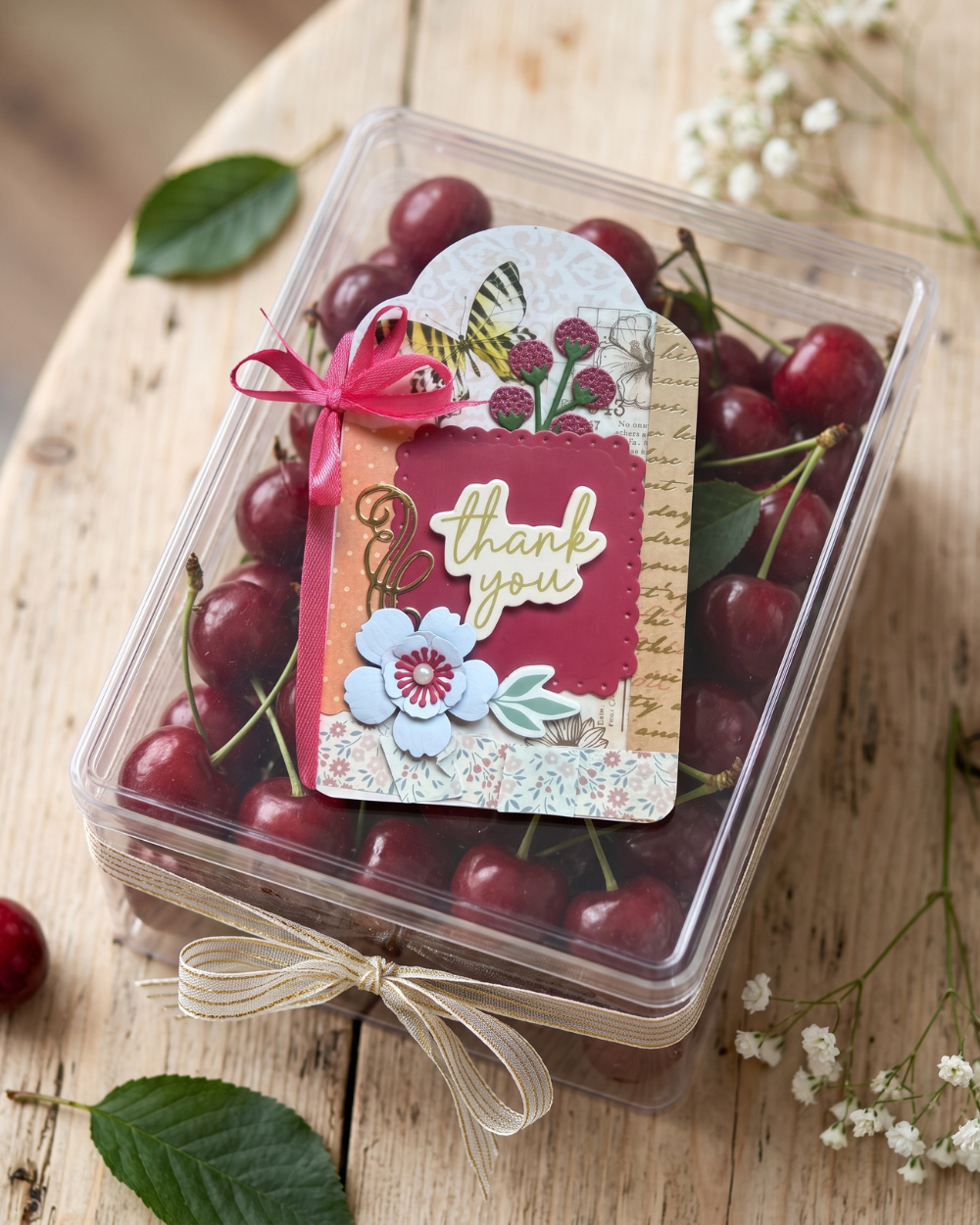 A clear plastic box filled with red cherries on a wooden surface, decorated with a handmade paper tag that says 'thank you' in gold script, tied with a pink ribbon and beige string.