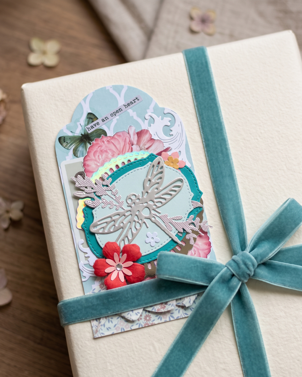 A close-up of a gift-wrapped package featuring a decorative layered paper tag with a dragonfly cutout, floral elements, and a teal ribbon tied in a bow on a cream fabric surface.