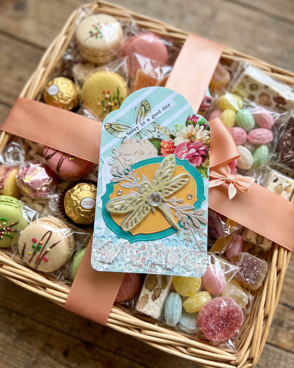 A wicker gift basket filled with assorted chocolates, macarons, and wrapped candies, tied with peach ribbons and topped with a decorative dragonfly card that reads 'Have a good day'.