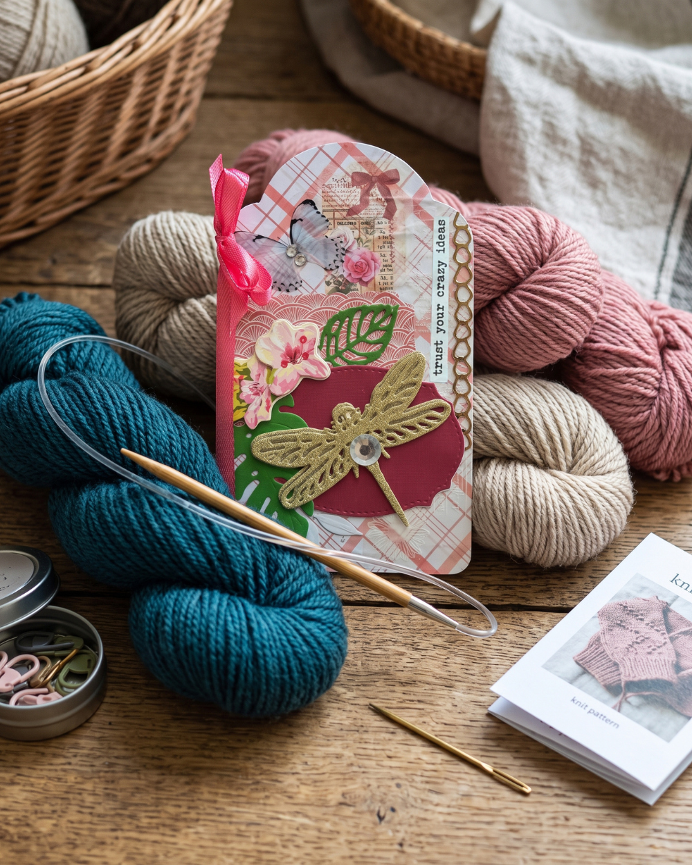 A knitted card featuring a dragonfly design and yarn information, styled on a wooden table with skeins of teal, pink, and beige yarn, knitting needles, a crochet hook, and a small booklet.