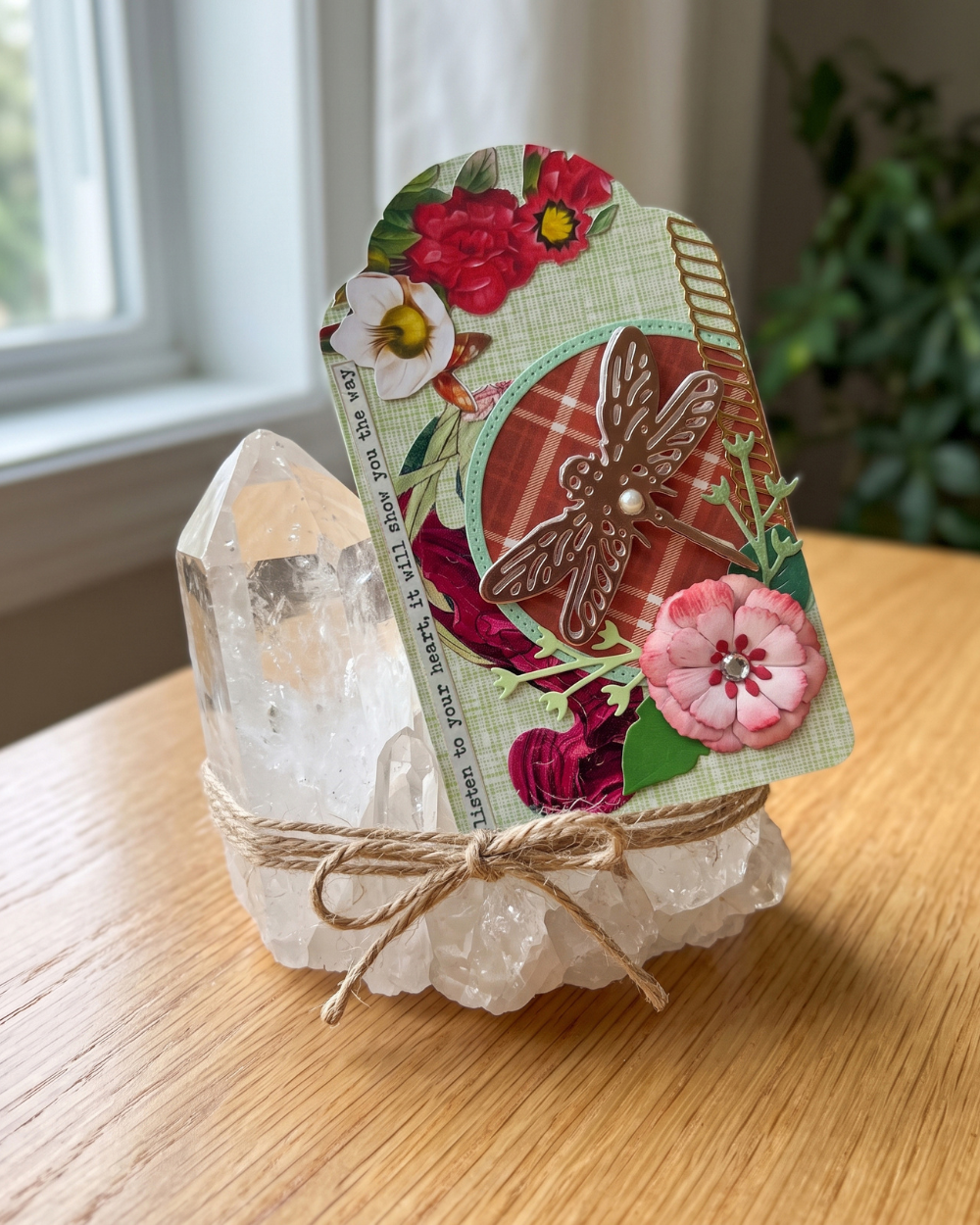 A decorative layered paper card with floral and dragonfly embellishments leaning against a clear crystal cluster wrapped in twine, placed on a wooden table near a window.