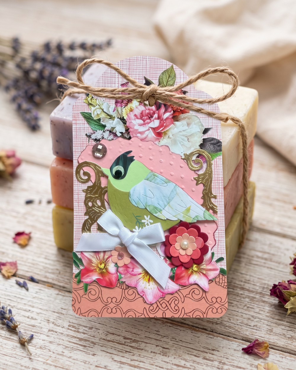 A decorative gift soap bar wrapped in twine with a floral paper tag featuring a green bird illustration, set on a rustic wooden surface with dried flowers and fabric in the background.